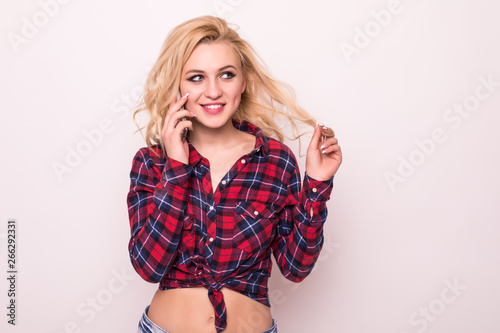 Thoughtful pretty young blonde woman in checkered shirt and jeans shorts talking on cell phone over white background © dianagrytsku