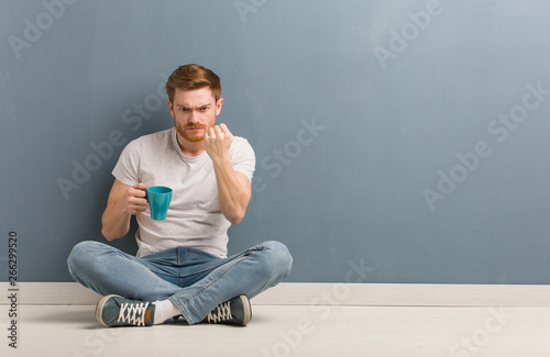 Young redhead student man sitting on the floor showing fist to front, angry expression. He is holding a coffee mug.