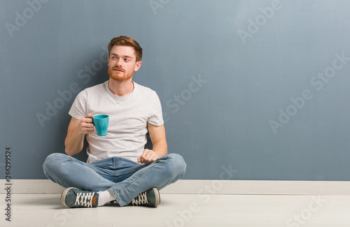 Young redhead student man sitting on the floor smiling confident and crossing arms, looking up. He is holding a coffee mug.