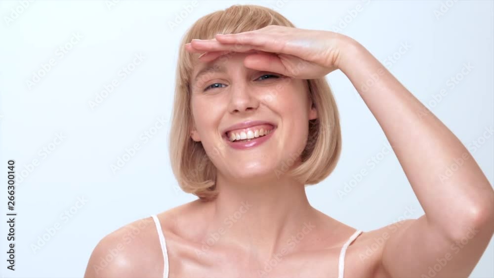 A beautiful young woman with blue eyes, blonde hair and a cute smile pretends to look in the sun for a beauty shoot in the studio on a white background.