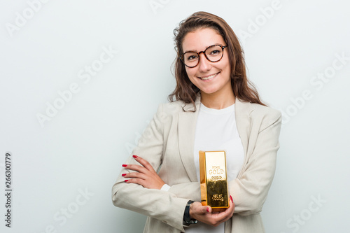 Fotografie Young european business woman holding a gold ingot smiling confident with crossed arms