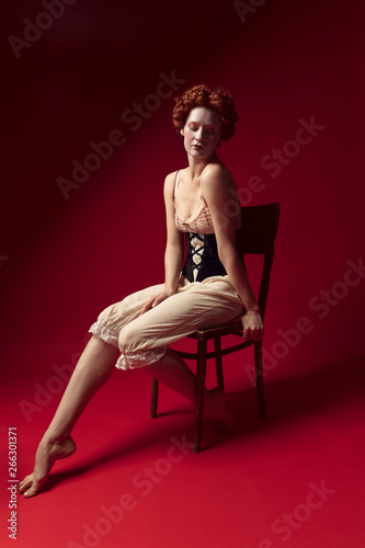 A little devilry. Medieval redhead young woman as a duchess in black corset and night clothes sitting on the chair on red background. Concept of comparison of eras, modernity and renaissance.