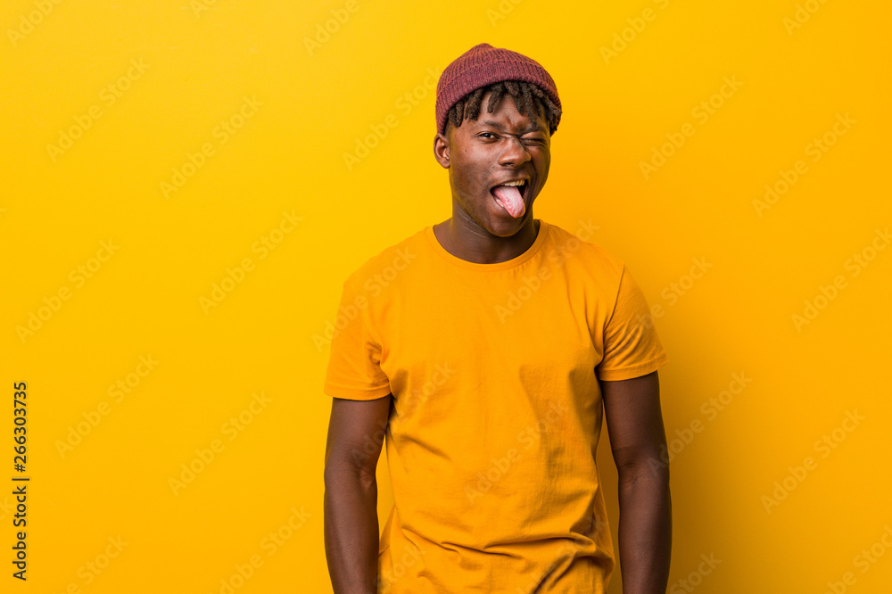 Young black man wearing rastas over yellow background funny and friendly sticking out him tongue.