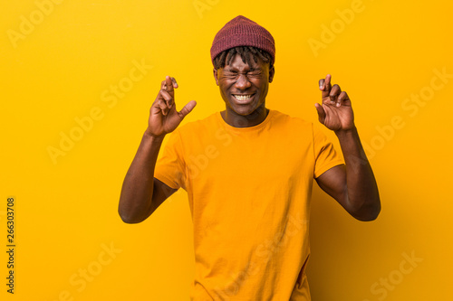 Young black man wearing rastas over yellow background crossing fingers for having luck