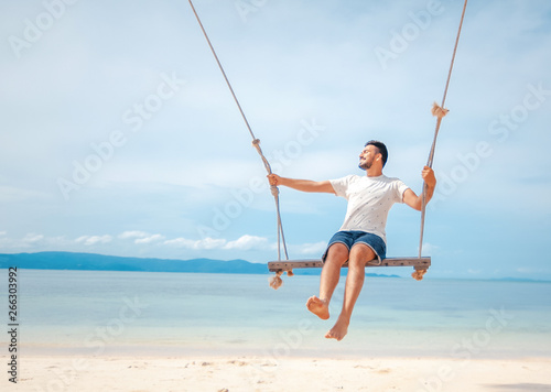 Young handsome attractive latin man rolling on a swing against the backdrop of a tropical beach and sea, vacations and travels