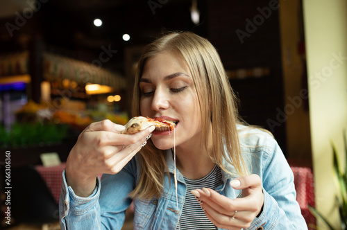 Funny blonde girl in jeans jacket eating pizza at restaurant. 