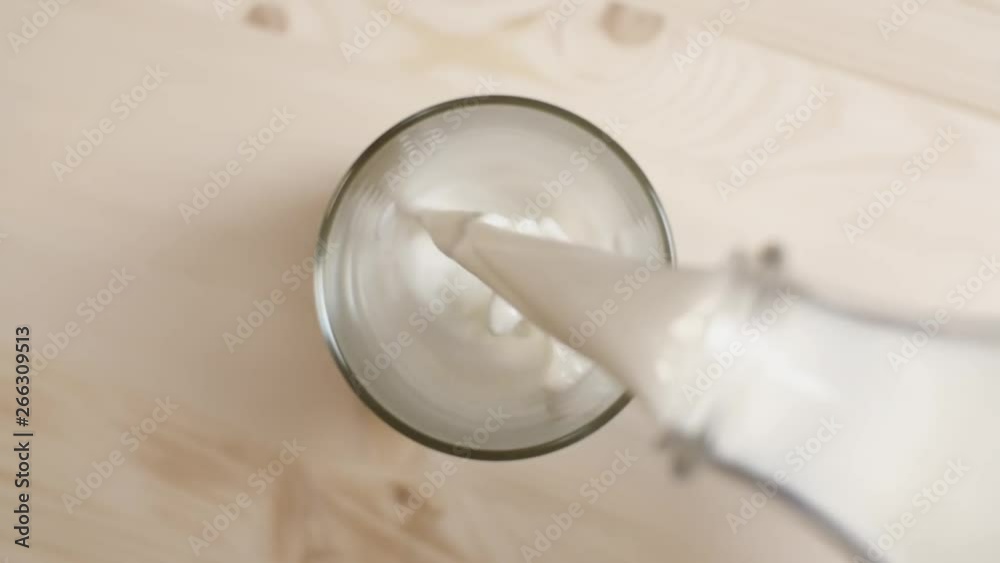 Top view of milk being poured in a glass on light brown wooden table, close up of milk pouring from transparent bottle on beige background, slow motion Top view of milk being poured in a glass on light brown wooden table, close up of milk pouring from transparent bottle on beige background, slow motion