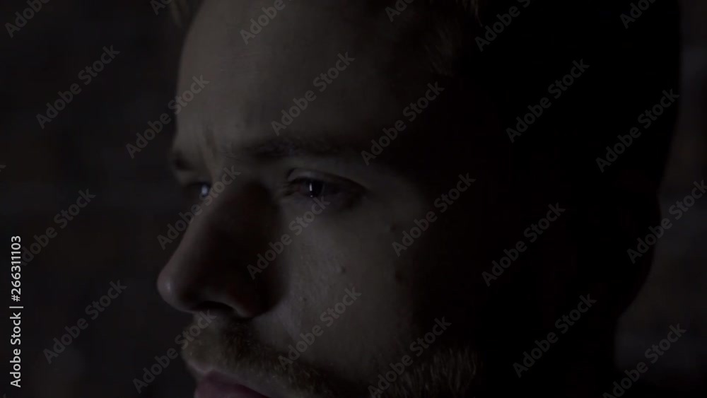 Very close up 4K of a handsome blond-haired young man with blue eyes looking away with a sad and frowning look and then looking at the camera with a light moving in a studio shed brick wall Very close up 4K of a handsome blond-haired young man with blue eyes looking away with a sad and frowning look and then looking at the camera with a light moving in a studio shed brick wall