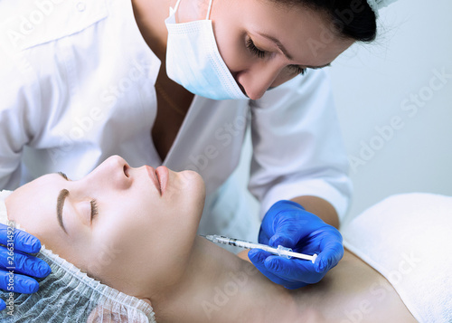 Beautician at work in beauty parlour in uniform outfit with face of patient and a disposable syringe in hand. Client lays on a couch with closed eyes and ready for procedure.