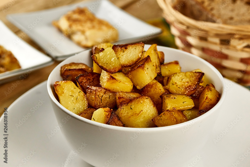 Roasted potatoes in a white bowl with rosemary and dill at next to fried onions and fish in batter on wooden background. Close-up rustic composition. Roasted potatoes in a white bowl with rosemary and dill at next to fried onions and fish in batter on wooden background. Close-up rustic composition.