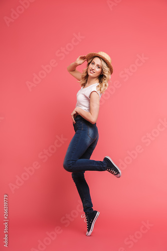 Fototapeta Naklejka Na Ścianę i Meble -  Full length portrait of positive blond woman 20s wearing casual jeans and straw hat smiling and walking