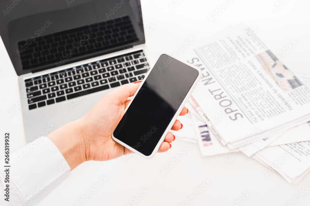 selective focus of woman holding smartphone with blank screen near laptop and newspapers with sport news selective focus of woman holding smartphone with blank screen near laptop and newspapers with sport news