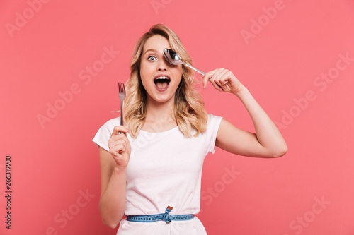 Portrait of excited blond woman 20s wearing body measuring tape around waist holding spoon and fork