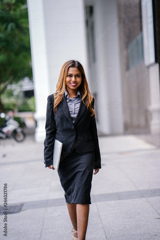 A beautiful and young Asian Malay woman is smiling confidently walking down a path by the street in the city during the day. She is professionally dressed and is holding her laptop. A beautiful and young Asian Malay woman is smiling confidently walking down a path by the street in the city during the day. She is professionally dressed and is holding her laptop.