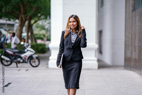 A beautiful and young Asian Malay woman is smiling confidently walking down a path by the street in the city during the day.  She is professionally dressed and is holding her laptop. © Danon