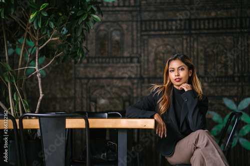 A sophisticated, young, tanned professional Southeast Asian business woman in a turtle neck top looking at the viewer and is sitting on a bench in the city restaurant during the day.