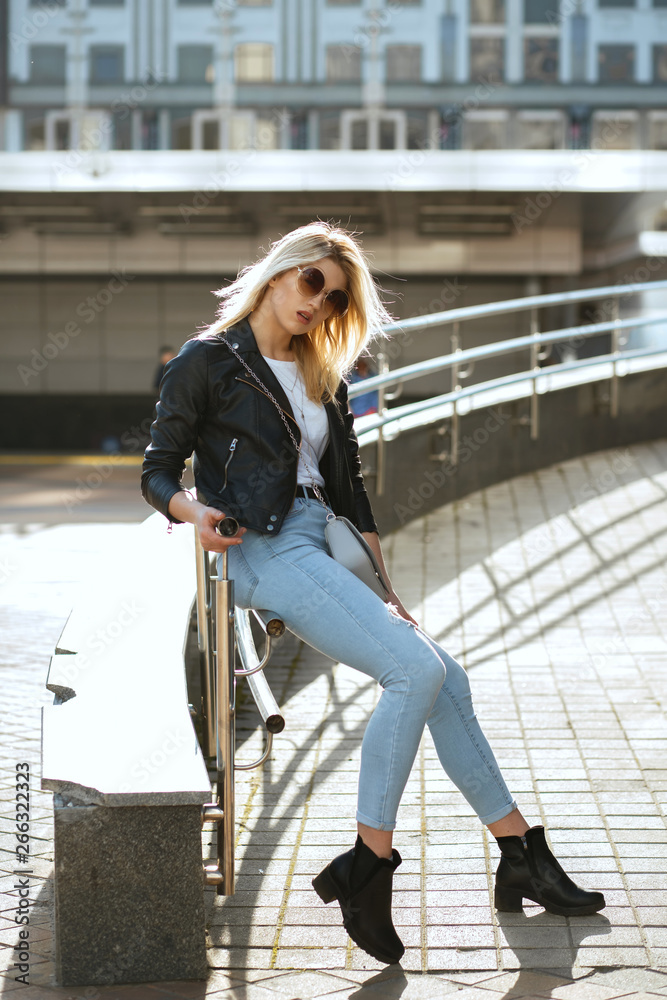 Full length shot of stylish blonde girl wearing vintage glasses sitting on street parapet
