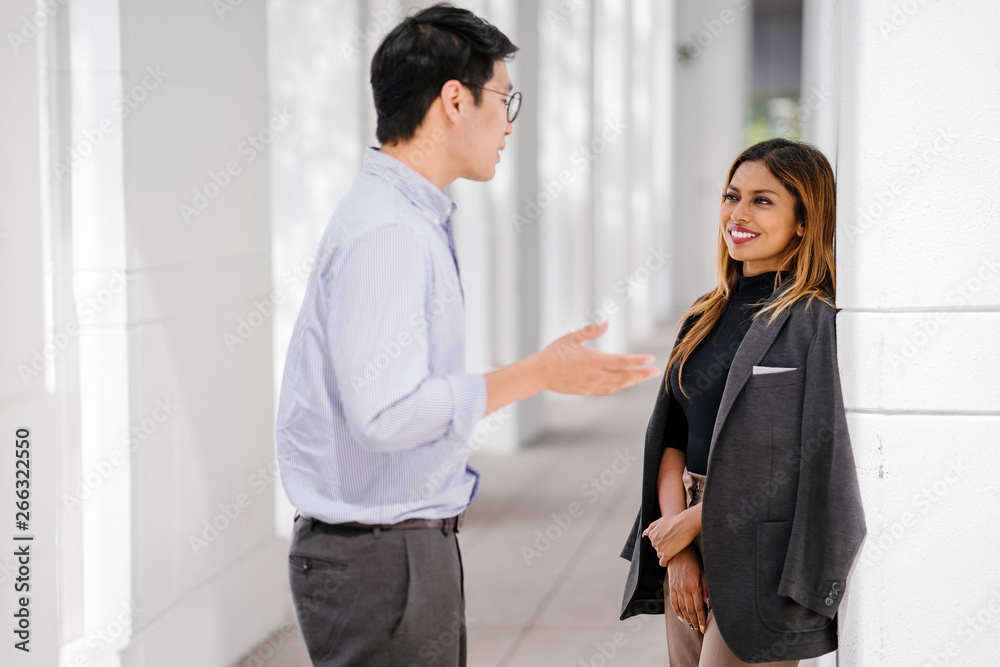 Candid shot of two diverse Asian business enthusiast, Korean man and a Malay woman having an animated conversation as they standby the hall during the day. They're both wearing a corporate attire. Candid shot of two diverse Asian business enthusiast, Korean man and a Malay woman having an animated conversation as they standby the hall during the day. They're both wearing a corporate attire.