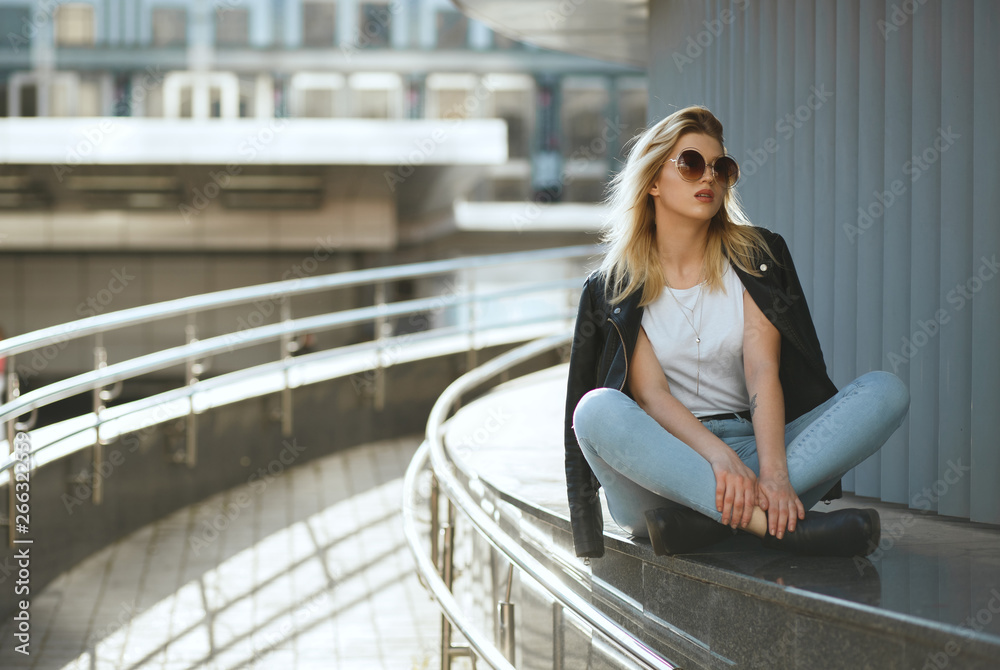 Amazing stylish woman wearing jeans and jacket sitting on street parapet with legs crossed. Copy space