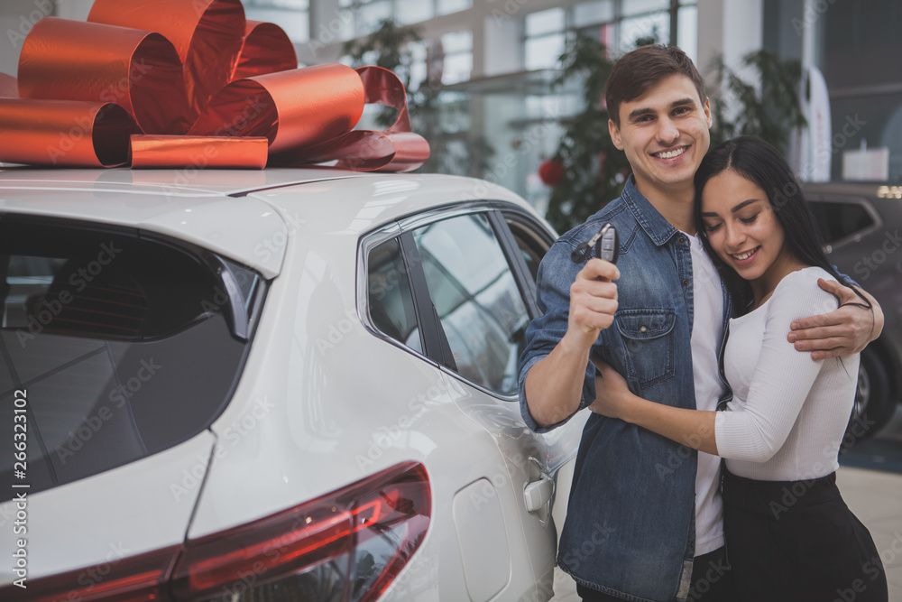 Handsome happy man smiling joyfully, hugging his gorgeous girlfriend after buying new car at the dealership. Lovely couple holding car keys, posing near their new automobile with red bow on the roof
