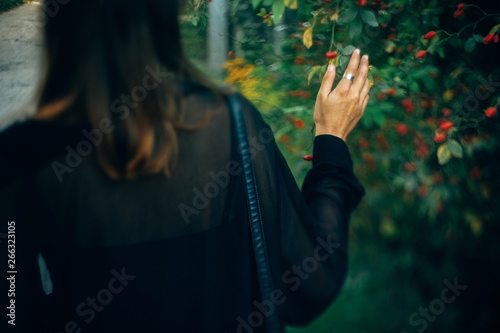 Stylish hipster girl walking in sunny light in park, atmospheric moment, back view. Fashionable woman holding branch at bush in evening. Selective focus. Space for text. Creative image