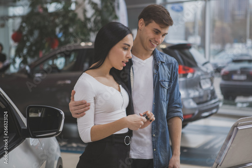 Beautiful happy couple reading information about new automobile on sale at the dealership salon. Handsome man hugging his beautiful wife, while choosing new car at the showroom. Buying car concept