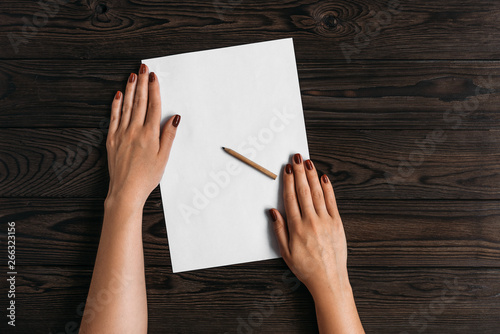 Top view of women's hands, ready to write something on an empty piece of paper lying on a wooden table. White blank sheet of paper with pencil on wooden background. Write a letter on a white sheet