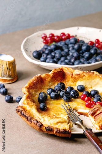 Started eaten fresh baked Dutch baby pancake in ceramic plate with blackberry and red currant berries, bowl of honey, jug of cream, vintage cutlery over beige kitchen table. Close up