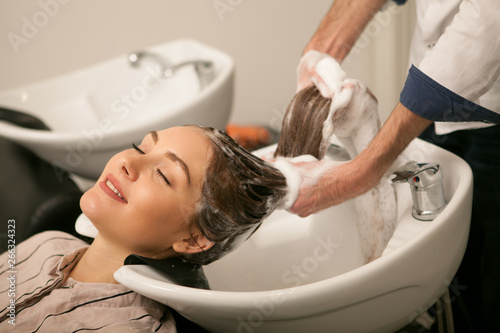 Attractive younf woman smiling with her eyes closed enjoying hair wash at beauty salon. Professional hairdresser washing hair of his female client, using shampoo. Cosmetics, hair care concept