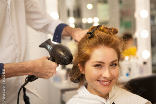 Happy young beautiful woman smiling to the camera excitedly, while hairdresser styling her long hair. Professional hairstylist working with female client, copy space. Blow drying, hairdo concept