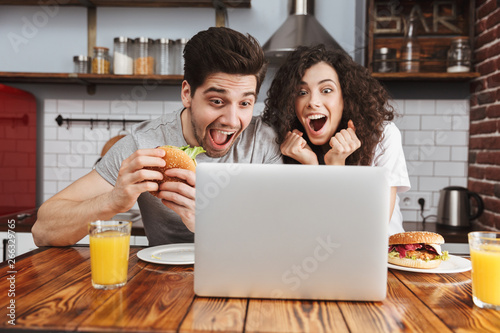 Picture of caucasian couple looking at laptop on table while eating hamburger in kitchen at home