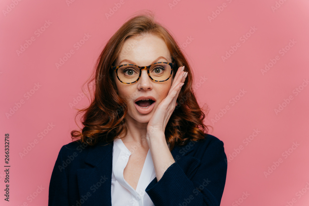 Really no way? Shocked red haired woman touches cheek, opens mouth widely, looks surprisingly at camera, finds out shocking news, wears spectacles, elegant formal suit, isolated on pink background Really no way? Shocked red haired woman touches cheek, opens mouth widely, looks surprisingly at camera, finds out shocking news, wears spectacles, elegant formal suit, isolated on pink background