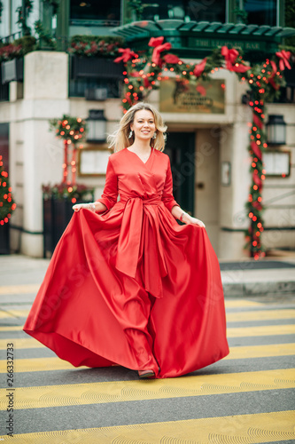 Christmas portrait of beautiful woman with blond hair, wearing long red dress, walking down the street