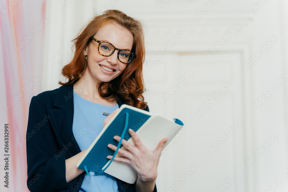 Prosperous satisfied businesswoman in elegant clothes writes in diary, has glad expression, wears spectacles, makes list of plans, writes text message in notebook, stands indoor. Female makes notes Prosperous satisfied businesswoman in elegant clothes writes in diary, has glad expression, wears spectacles, makes list of plans, writes text message in notebook, stands indoor. Female makes notes