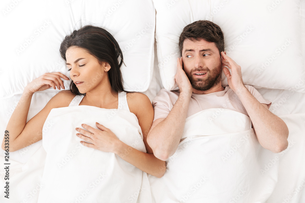Displeased man covering ears near his woman sleeping in bed under blanket.