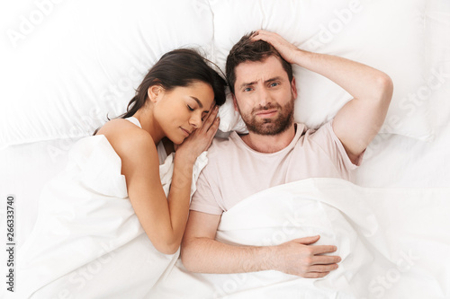 Displeased confused young man lies in bed under blanket near sleeping woman.
