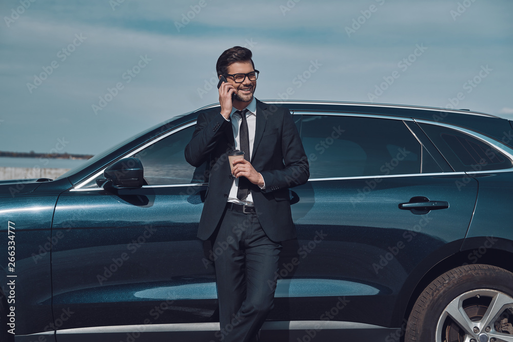 I am waiting for you! Handsome young businessman talking on his smart phone and smiling while standing near his car outdoors I am waiting for you! Handsome young businessman talking on his smart phone and smiling while standing near his car outdoors
