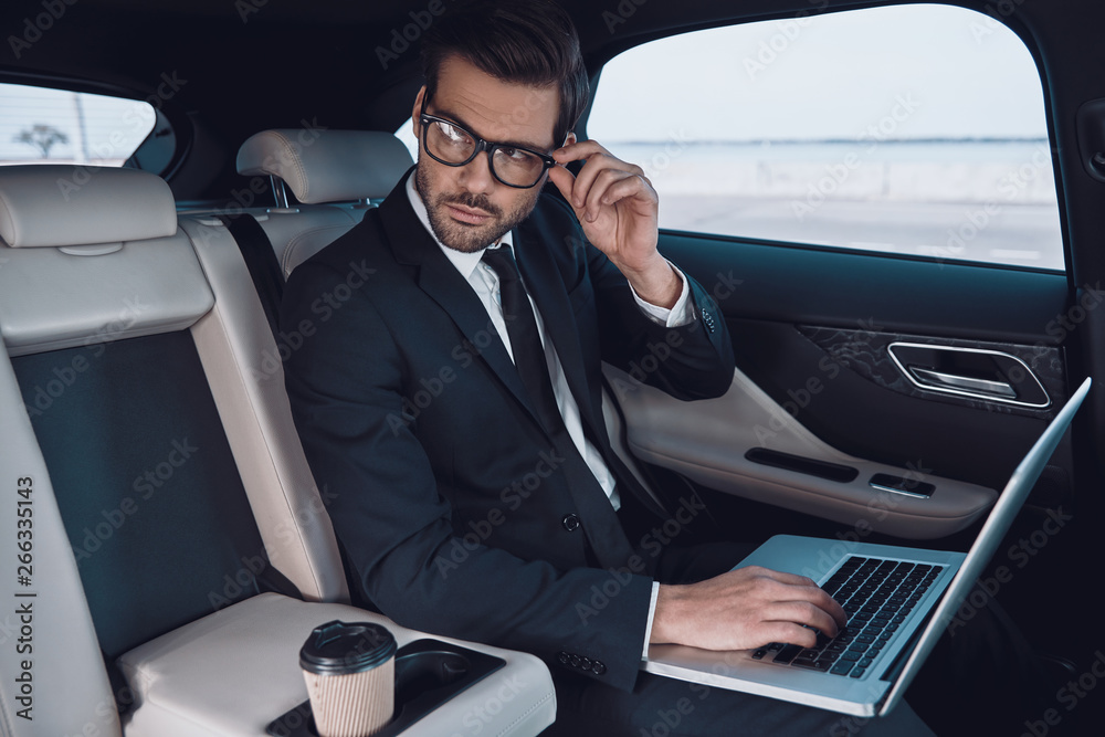 Planning his business. Thoughtful young man in full suit working using laptop and adjusting his eyewear while sitting in the car