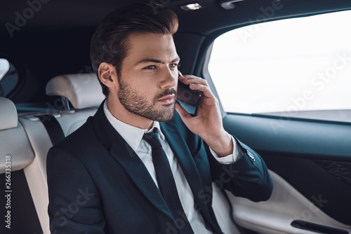 Taking everything seriously. Handsome young man in full suit talking on the smart phone while sitting in the car