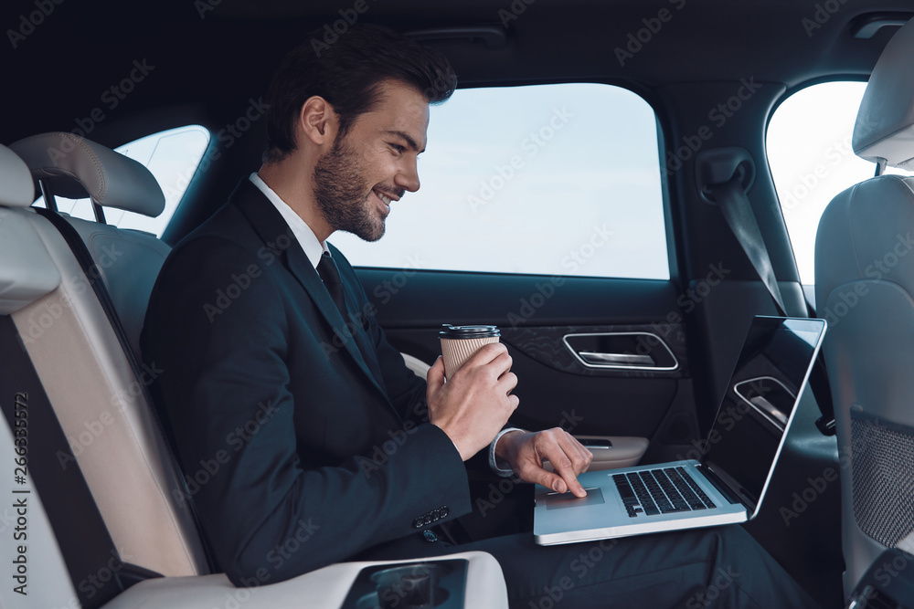 Driven by success. Handsome young man in full suit working using laptop while sitting in the car Driven by success. Handsome young man in full suit working using laptop while sitting in the car