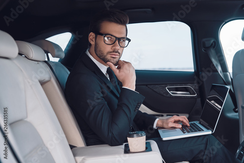 Successful professional. Thoughtful young man in full suit working using laptop and adjusting his eyewear while sitting in the car