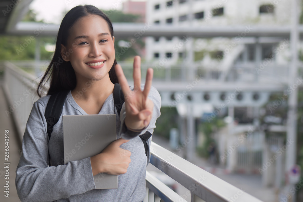portrait of happy smiling asian woman college student pointing up 3 fingers, three points pose; winning, third concept in city campus environment
