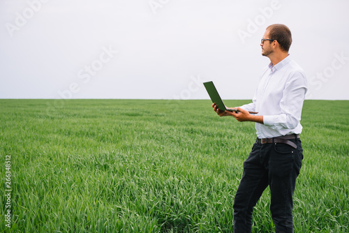 Young agronomist holds laptop in green wheat field. Agribusiness concept. agricultural engineer standing in a wheat field with a tablet in summer