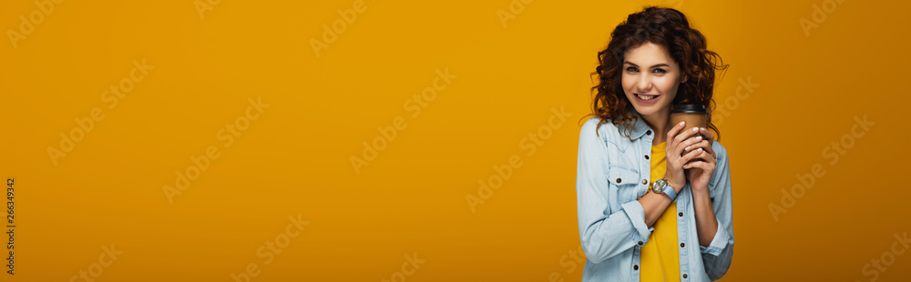 panoramic shot of curly redhead girl holding paper cup while smiling  on orange