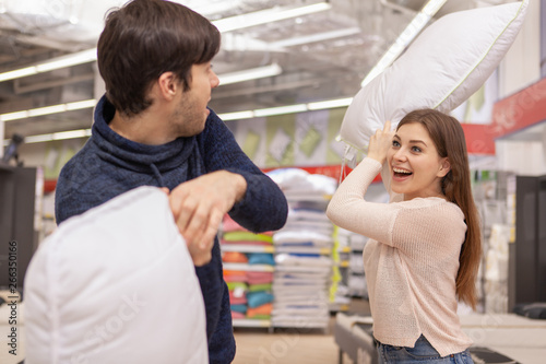 Excited beautiful woman screaming cheerfully, fighting her boyfriend with a pillow at furniture store. Loving happy couple having pillowfight while shopping home goods. Love, couples, valentine concep