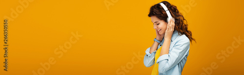 panoramic shot of happy redhead girl listening music in headphones on orange