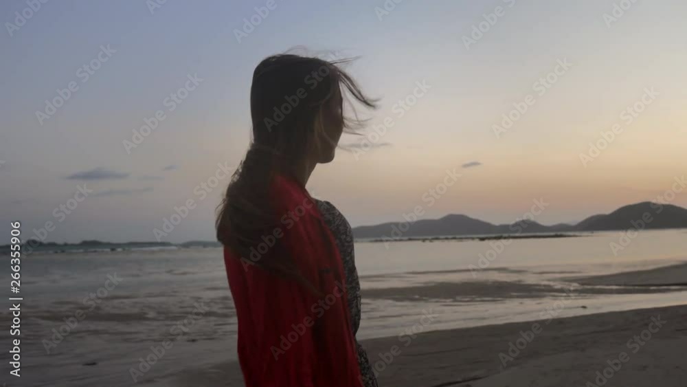 Young attractive brunette woman model walks in sunset on the tropical beach 4k. Red cape and dress. Thoughtful beautiful girl in golden hour. Wind blows her hair. Camera slides around the person Young attractive brunette woman model walks in sunset on the tropical beach 4k. Red cape and dress. Thoughtful beautiful girl in golden hour. Wind blows her hair. Camera slides around the person