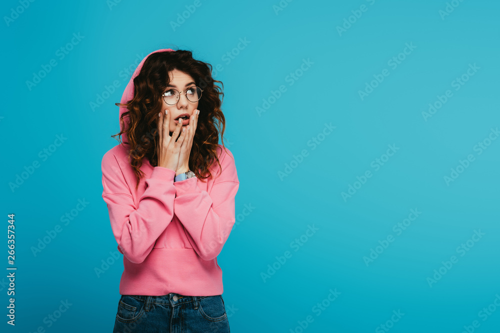surprised redhead girl touching face while standing on blue
