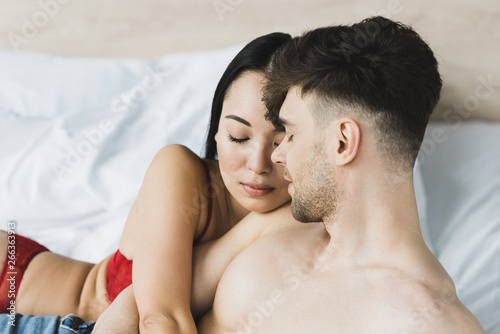beautiful asian woman in red lingerie hugging shirtless boyfriend while lying in bed together