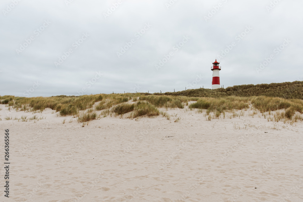 White - red lighthouse at the beach with sand and grass dunes, cloudy sky
